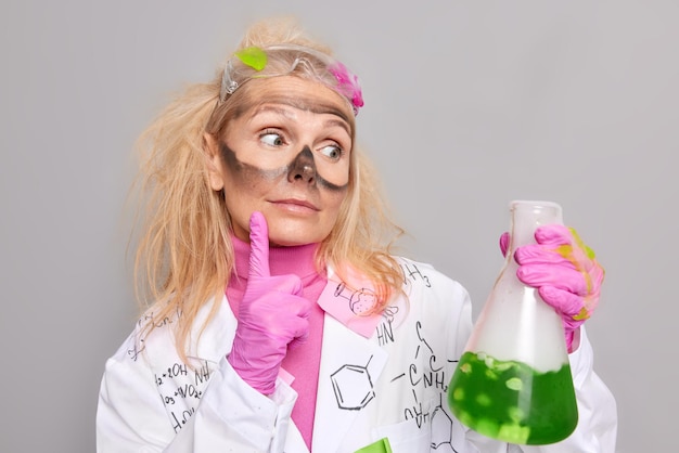 Der Chemiker sieht sich aufmerksam Glasbecher mit grüner Flüssigkeit an, die im Chemielabor arbeiten, trägt Gummihandschuhe und einen medizinischen Mantel einzeln auf grauem Hintergrund. Wissenschafts- und Forschungskonzept