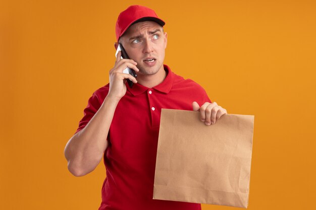 Der denkende Blick auf den jungen Lieferboten der Seite, der Uniform mit Kappe hält, die Papiernahrungsmittelpaket hält, spricht am Telefon lokalisiert auf orange Wand