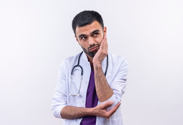 Der denkende junge männliche Arzt, der das medizinische Kleid des Stethoskops trägt, legte seine Hand auf Wange auf isoliertem Weiß