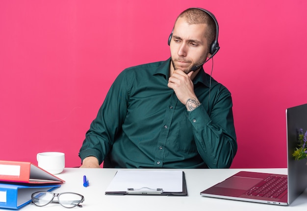 Der denkende junge männliche Callcenter-Betreiber, der ein Headset am Schreibtisch mit Bürowerkzeugen trägt, packte das Kinn