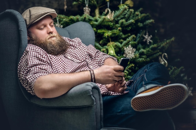 Der dicke bärtige Mann sitzt auf einem Stuhl und benutzt ein Smartphone mit einem Weihnachtsbaum im Hintergrund.