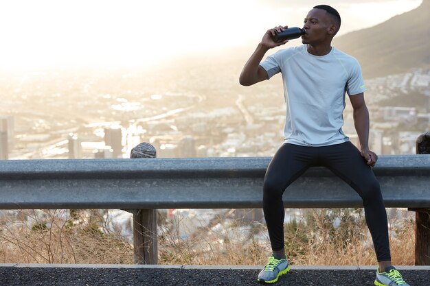 Der durstige Afroamerikaner trinkt frisches Wasser, genießt eine Pause nach dem Sporttraining unter freiem Himmel und sitzt am Straßenschild mit dem malerischen Panoramablick auf die Berge für Werbeinhalte oder Informationen