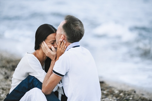 Der Ehemann küsst zärtlich seine schöne Frau am Strand in der Nähe des stürmischen Meeres in die Stirn und lächelt