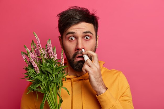 Der empörte bärtige Mann mit den wässrigen Augen besprüht die Nase mit Tropfen, fühlt sich wegen einer Allergie krank, trägt ein gelbes Sweatshirt, behandelt saisonale Krankheiten, ist auf einer rosa Wand isoliert und hat Rötungen um die Augen