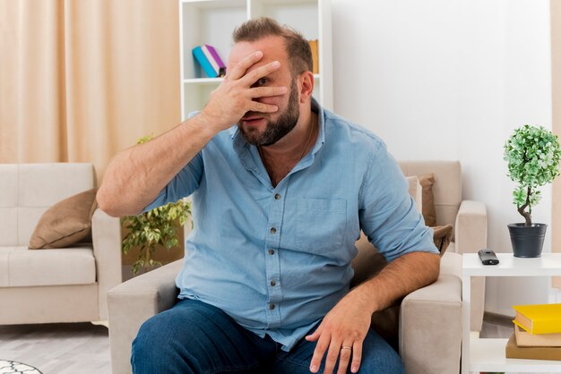 Der enttäuschte erwachsene slawische Mann sitzt auf einem Sessel und legt die Hand auf das Gesicht, wobei er die Kamera durch die Finger im Wohnzimmer betrachtet