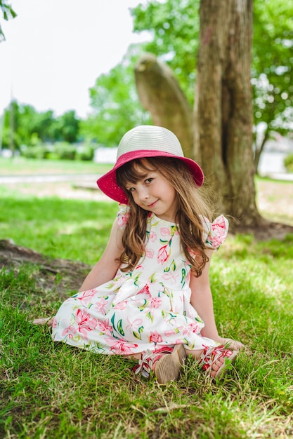 Der entzückende kleine Mädchen auf dem Boden mit einem Hut sitzt in einem Park