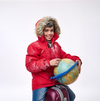 Der entzückende schuljunge in einem warmen, schneebedeckten roten parka mit kapuze, der auf einem koffer sitzt, lächelt zähnefletschend und zeigt mit dem finger auf den globus auf der suche nach einem reiseziel. wintertourismuskonzept