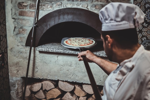 Kostenloses Foto der erfahrene koch bringt seine pizza mit einem speziellen riesenspatel in den ofen.