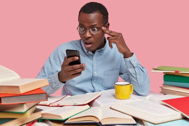 Der erstaunte schwarze Mann starrt auf das Handy, liest Nachrichten auf der Internet-Website, trägt formelle Kleidung und bereitet sich allein auf das Seminar vor
