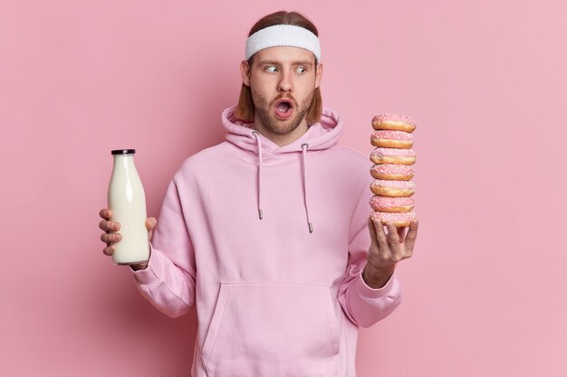 Der erstaunte sportliche Mann starrt auf leckere, köstliche Donuts und hat die Versuchung, in einem weißen Stirnband mit Kapuzenpulli zu essen.