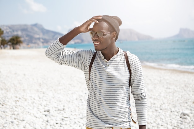 Der fröhliche afrikanische junge Mann, der am Strand stand und mit einem entzückenden Lächeln zur Seite schaute, bemerkte, dass sein Freund kam und den Kopf berührte. Sommerferien und Abenteuer. Menschen, Lebensstil und Reisen