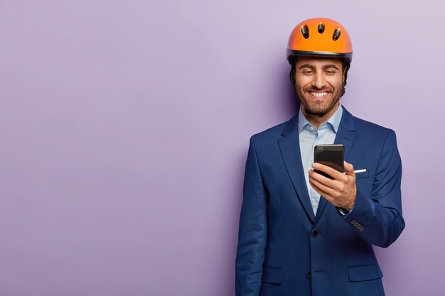 Der fröhliche Architekt trägt einen orangefarbenen Helm, einen formellen Anzug, hat während der Arbeitspause Freizeit, erhält eine Nachricht auf dem Smartphone und freut sich über das Gehalt