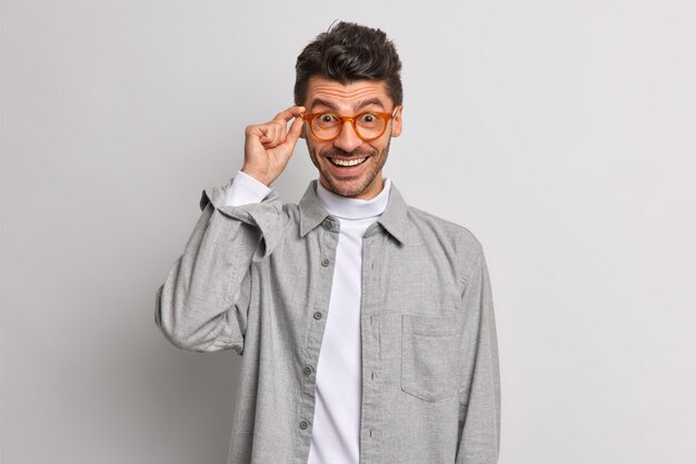 Der fröhliche, gutaussehende europäische Kerl sieht glücklich durch eine Brille aus, die in grauem Hemd gekleidet ist und lächelt unbeschwerte Posen im Innenbereich. Glücklicher männlicher Angestellter bereit für die nächste Aufgabe. Konzept der positiven Emotionen