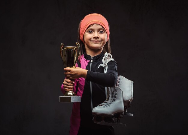 Der glückliche kleine Skater-Mädchen-Champion hält einen Siegerpokal und Schlittschuhe. Getrennt auf einem dunklen strukturierten Hintergrund.