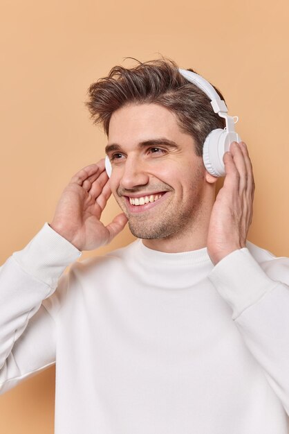 Der gutaussehende, fröhliche europäische Mann lächelt angenehm und hält die drahtlosen Stereo-Kopfhörer in den Händen. Hobbykonzept