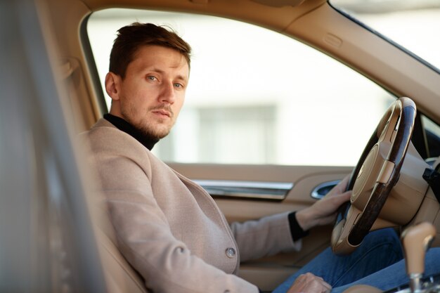 Der gutaussehende kaukasische Fahrer in beiger Jacke sitzt auf dem Vordersitz eines neuen Autos und hält das Lenkrad
