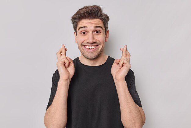 Der hoffnungsvolle, fröhliche europäische erwachsene Mann macht den Wunsch zuversichtlich, ein Lächeln zu gewinnen, trägt glücklich ein lässiges schwarzes T-Shirt und drückt die Daumen, dass er an Glück glaubt, oder dass das Glück kommen wird. Konzept der Körpersprache