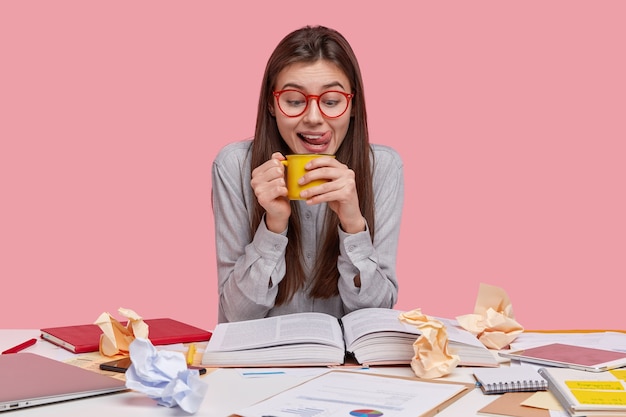 Der horizontale Schuss der Studentin hält eine Tasse mit einem aromatischen Getränk, leckt sich die Lippen, trägt eine Brille und ein Hemd, liest ein Buch und bereitet sich auf die Prüfung vor