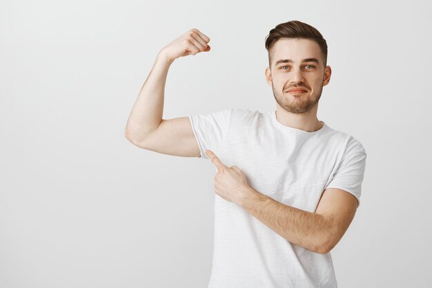 Der hübsche junge Mann zeigt seine Kraft, wenn er ins Fitnessstudio geht, um zu trainieren, Muskeln zu spielen und stolz zu lächeln