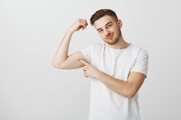 Der hübsche junge Mann zeigt seine Kraft, wenn er ins Fitnessstudio geht, um zu trainieren, Muskeln zu spielen und stolz zu lächeln