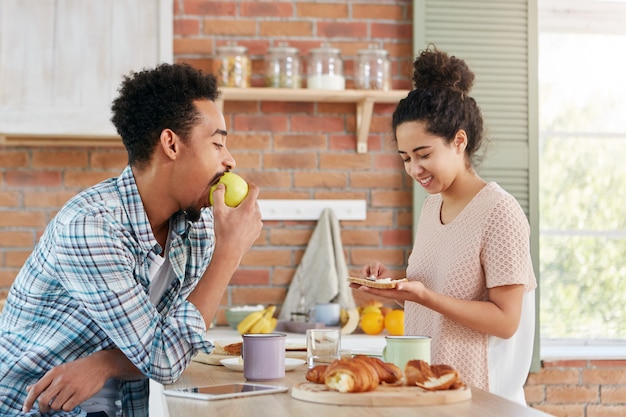 Der hungrige junge Mann gemischter Rassen isst Apfel, während er wartet, wenn seine Frau das Abendessen kocht. Lockige schöne Frau macht Schlangen