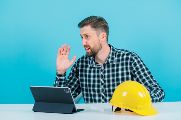 Kostenloses Foto der ingenieur schreit, indem er die hand in der nähe des mundes hält und vor der tablet-kamera auf blauem hintergrund sitzt