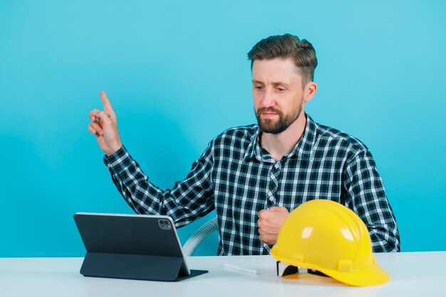 Der Ingenieurmann posiert für die Tablet-Kamera, indem er mit dem Zeigefinger auf blauem Hintergrund nach oben zeigt