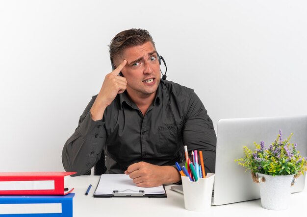 Der junge ängstliche blonde Büroangestellte Mann auf Kopfhörern sitzt am Schreibtisch mit Bürowerkzeugen unter Verwendung des Laptops setzt Finger auf Kopf, der Kamera lokalisiert auf weißem Hintergrund mit Kopienraum betrachtet