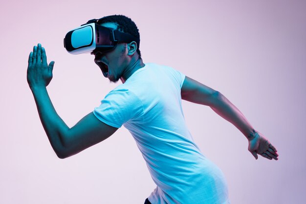 Der junge Afroamerikaner spielt und benutzt VR-Brille im Neonlicht auf Gradient