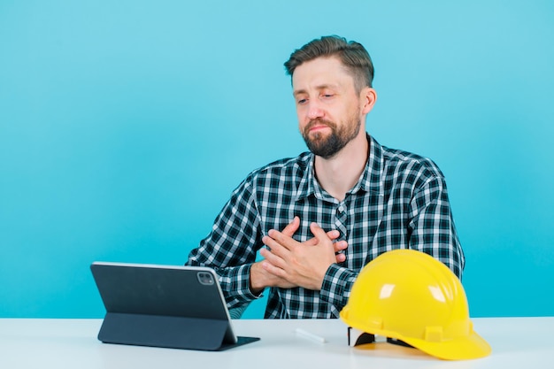 Der junge Architekt hält die Hände am Herzen, indem er vor dem Tablet auf blauem Hintergrund sitzt