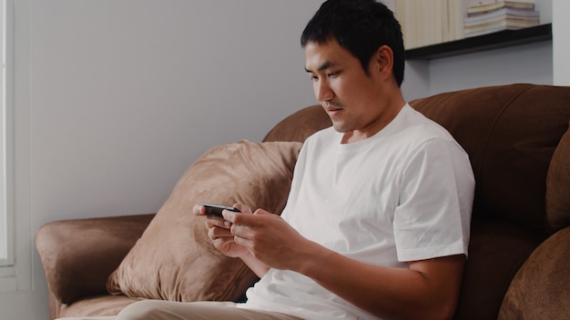 Der junge asiatische Mann, der den Handy spielt Videospiele im Fernsehen im Wohnzimmer verwendet, der Mann, der unter Verwendung glücklich sich fühlt, entspannen sich die Zeit, die zu Hause auf Sofa liegt. Männer spielen zu Hause entspannen.