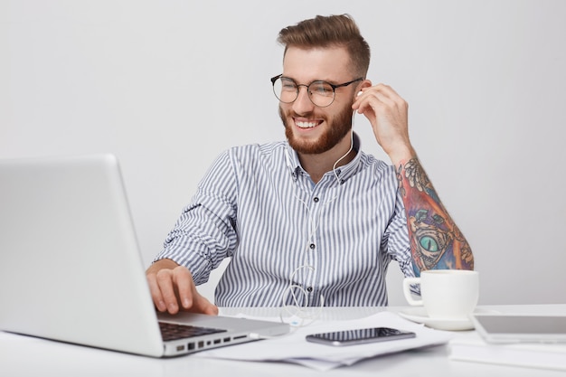 Der junge attraktive Geschäftsmann hat einen glücklichen Ausdruck, wenn er sich nach harter Arbeit ausruht, Musik hört oder Filme mit Kopfhörern und Laptop ansieht. Tätowierter Hipster-Student genießt Lieblings-Audio-Trac