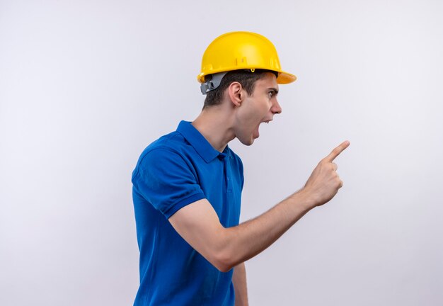 Der junge Baumeister, der die Bauuniform und den Schutzhelm trägt, zeigt wütend mit dem Zeigefinger nach links