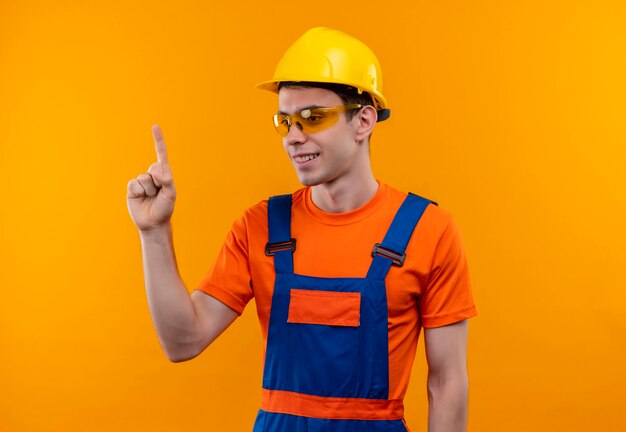 Der junge Baumeister, der eine Schutzbrille und einen Schutzhelm trägt, lächelt und zeigt mit den Daumen nach oben