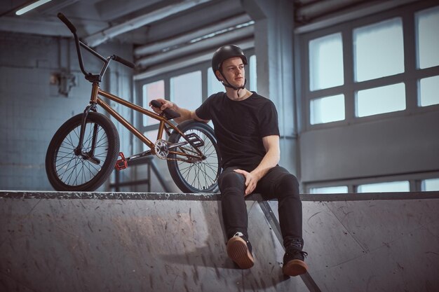 Der junge Bmx-Fahrer entspannt sich, nachdem er Tricks geübt hat, die sich auf sein Fahrrad lehnen und in einem Skatepark drinnen wegschauen