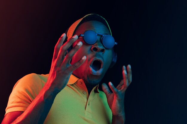 Der junge gutaussehende glückliche überraschte Hipster-Mann, der Musik mit Kopfhörern im schwarzen Studio mit Neonlichtern hört. Disco, Nachtclub, Hip-Hop-Stil, positive Emotionen, Gesichtsausdruck, Tanzkonzept
