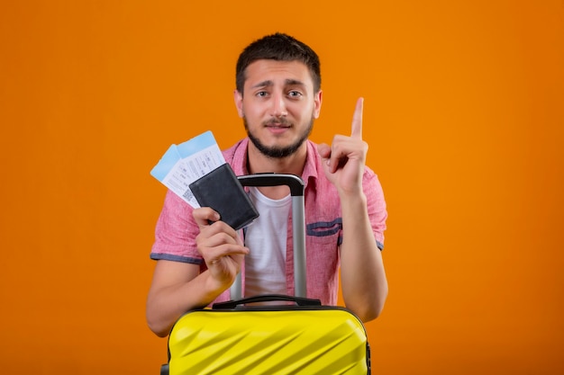 Der junge gutaussehende Reisende, der Flugtickets hält, die mit dem Finger nach oben zeigen, erinnert sich daran, wichtige Dinge nicht zu vergessen, die mit Koffer über orange Hintergrund stehen