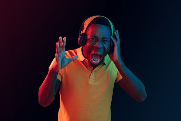 Der junge hübsche glückliche Hipster-Mann, der Musik mit Kopfhörern im schwarzen Studio mit Neonlichtern hört. Disco, Nachtclub, Hip-Hop-Stil, positive Emotionen, Gesichtsausdruck, Tanzkonzept