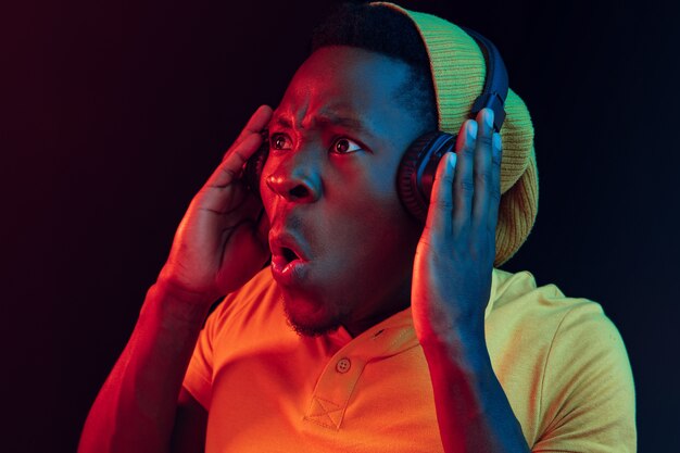 Der junge hübsche glückliche Hipster-Mann, der Musik mit Kopfhörern im schwarzen Studio mit Neonlichtern hört. Disco, Nachtclub, Hip-Hop-Stil, positive Emotionen, Gesichtsausdruck, Tanzkonzept