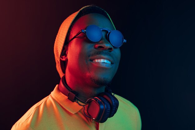 Der junge hübsche glückliche Hipster-Mann, der Musik mit Kopfhörern im schwarzen Studio mit Neonlichtern hört