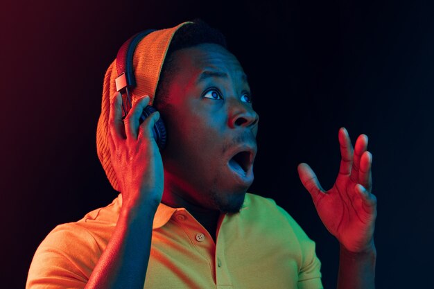 Der junge hübsche glückliche überraschte Hipster-Mann, der Musik mit Kopfhörern im schwarzen Studio mit Neonlichtern hört. Disco, Nachtclub, Hip-Hop-Stil, positive Emotionen, Gesichtsausdruck, Tanzkonzept