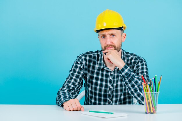 Der junge Ingenieur schaut in die Kamera, indem er die Hand am Kinn auf blauem Hintergrund hält