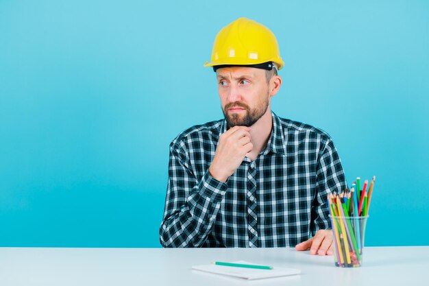 Der junge Ingenieur schaut nach links, indem er die Hand am Kinn auf blauem Hintergrund hält