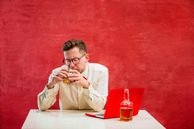 Der junge lustige Mann mit Cognac, der mit Laptop am Valentinstag auf rotem Studio sitzt.