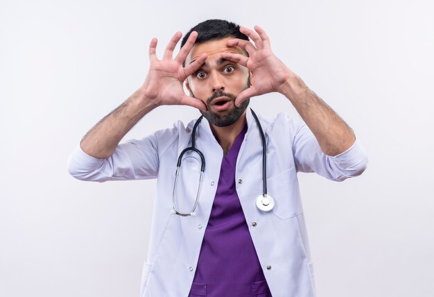 Der junge männliche Arzt, der ein medizinisches Stethoskopkleid trug, legte seine Hände um die Augen auf die isolierte weiße Wand