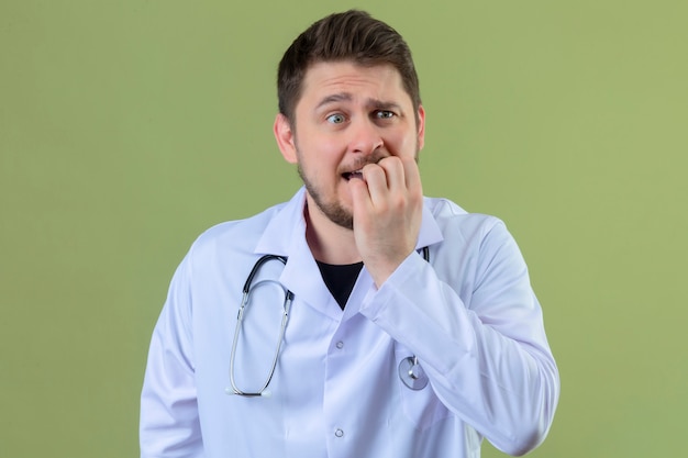 Der junge Mann Arzt, der weißen Kittel und Stethoskop trägt, hält Hände nahe Mund, fühlt sich erschrocken und erschrocken über isoliertem grünem Hintergrund