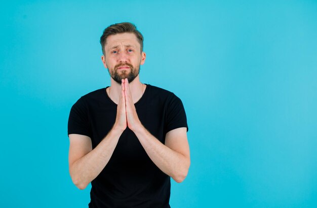 Der junge Mann bettelt, indem er die Hand auf der Brust auf blauem Hintergrund zusammenhält