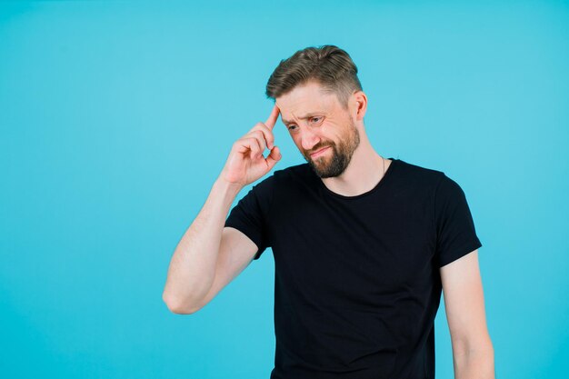 Der junge Mann denkt, indem er den Zeigefinger auf die Stirn hält und auf blauen Hintergrund blickt