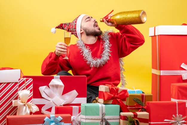 Der junge Mann der Vorderansicht, der herum sitzt, präsentiert Geschenke, die mit Champagner auf gelb feiern