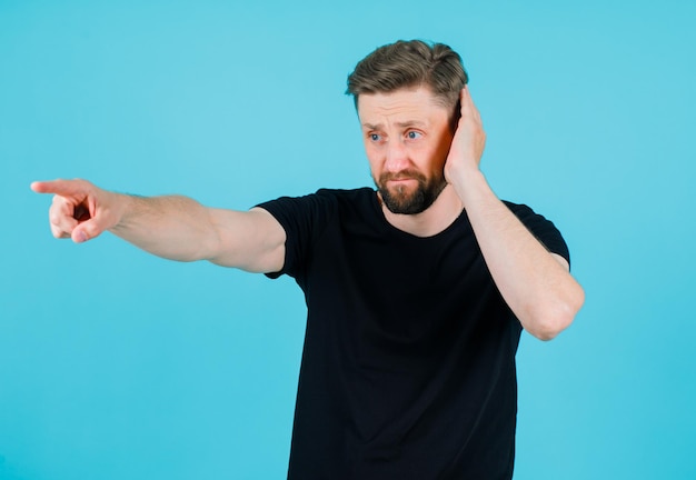 Kostenloses Foto der junge mann hält die hand am ohr und zeigt mit dem zeigefinger auf blauem hintergrund nach links